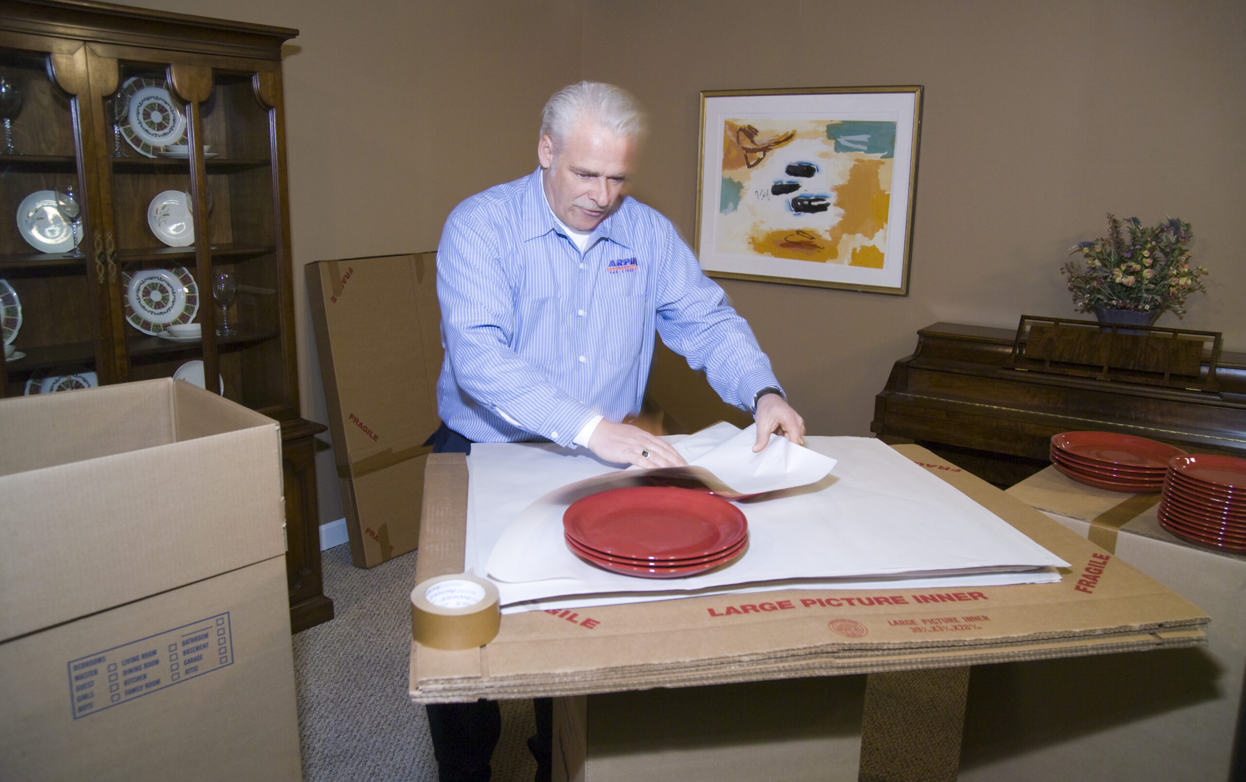 Arpin of RI packer in a dining room carefully wrapping fragile china plates with white newsprint.
